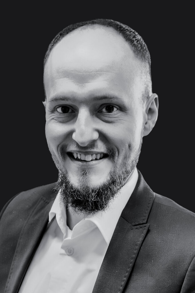 Ein Mann mit gestutztem Bart und kurzen Haaren, der eine Anzugsjacke und ein weißes Hemd trägt, lächelt in die Kamera vor einem schlichten dunklen Hintergrund.